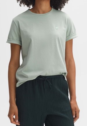 Vrouw draagt een lichtgroen kortärmelig t-shirt met een klein hartembleem op de borst en zwarte, getextureerde broek met trekkoord.