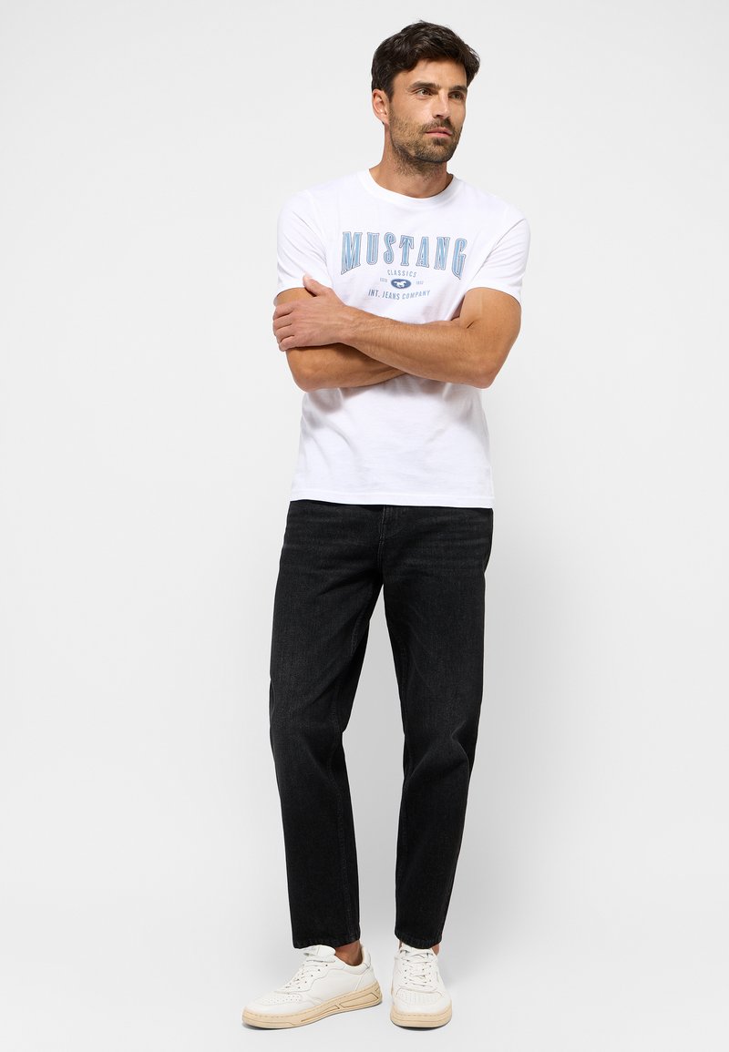 Mann, der mit verschränkten Armen auf einem einfarbigen Hintergrund steht, trägt ein weißes T-Shirt mit Mustang-Logo, schwarze Jeans und weiße Sneakers.