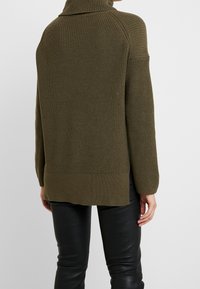 Maglione a maglia verde oliva con colletto alto, texture a coste, spalle scese e un orlo posteriore più lungo, abbinato a pantaloni neri aderenti.