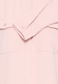 Blusa rosa chiaro con maniche lunghe, dotata di due tasche sul petto, chiusure con bottoni e una texture di tessuto liscia.