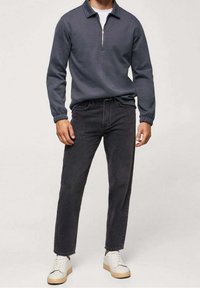 Homme portant un pull zippé gris foncé, un jean noir à jambe droite et des baskets blanches, debout devant un fond uni.