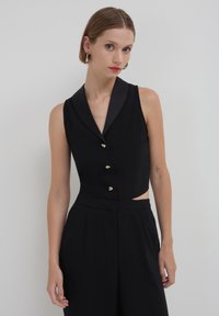 Combinaison noire sans manches avec col, dotée de boutons dorés et d'une découpe sur le côté. Le tissu a une texture lisse et un ajustement tailleur.