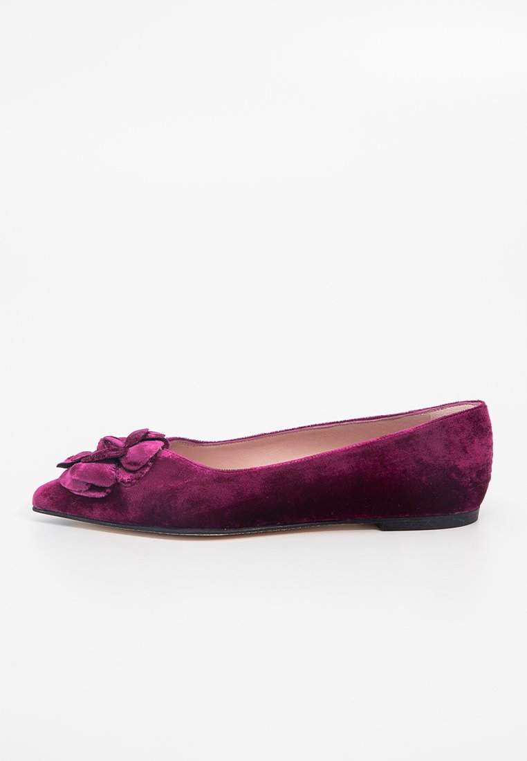 Zapato plano de ante fucsia con punta afilada, que cuenta con un adorno floral en la parte superior y una suela negra baja.