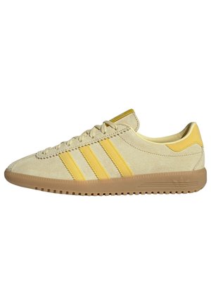 Beżowy, niskoprofilowy zamszowy sneaker z żółtymi paskami, żółtą pętelką na pięcie, gumową podeszwą i sznurowaniem.