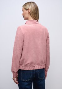 Kvinde med lige blont hår iført en ribbet lyserød jakke og blå jeans, set bagfra mod en ensfarvet lys baggrund.