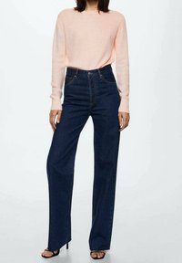 Blaue High-Waist-Jeans mit geradem Bein, kombiniert mit einem hellrosa Langarmpullover, getragen mit schwarzen Riemchensandalen.