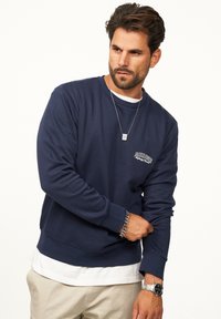 Navy blue sweatshirt in soft fabric, featuring a crew neck, long sleeves, and a small white logo on the left chest. Layered over a white shirt.