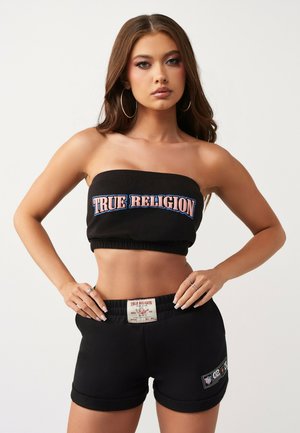 Młoda kobieta w czarnym, bezramkowym crop topie i pasujących szortach True Religion stoi, trzymając ręce na biodrach, na białym tle.