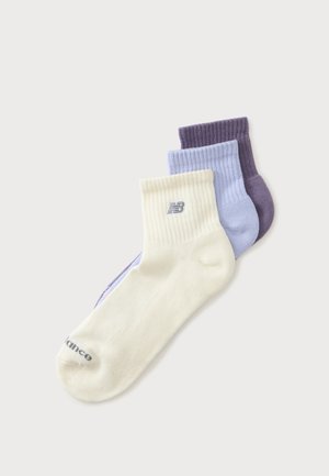 Trois paires de chaussettes côtelées de couleur crème, bleu clair et violet foncé, empilées avec un logo New Balance visible sur la paire crème.