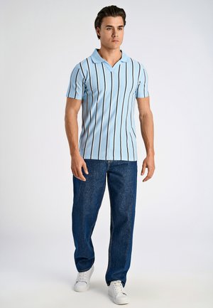 Hellblaues Poloshirt mit kurzen Ärmeln und schwarzen vertikalen Streifen, kombiniert mit dunklen Jeans und weißen Sneakers. Einfarbiger Hintergrund.