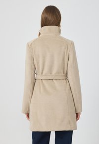 Beige takki, joka on valmistettu pehmeästä kankaasta, jossa on korkea kaulus, muotoiltu vyötärö vyöllä ja suora leikkaus.