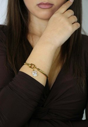 Mujer con una camiseta de manga larga de color marrón oscuro, mostrando una pulsera de oro con un charm cuadrado de piedra transparente en su muñeca izquierda.