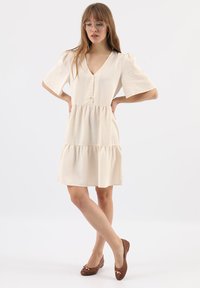 Cremefarbenes Kleid mit kurzen Puffärmelchen, V-Ausschnitt und Knopfdetails vorne. Gestuftes Rüschensaum und glatte Textur. Braune Ballerinas mit Schleifenakzenten.