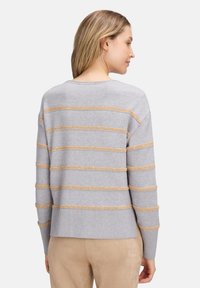 Frau mit blonden Haaren, die einen hellgrauen Pullover mit beigen horizontalen Streifen und beigen Hosen trägt, von hinten gegen einen schlichten Hintergrund gerichtet.