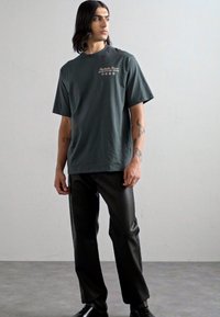 Camiseta de algodón de color verde oscuro con mangas cortas y escote redondo, que presenta un texto gráfico claro. Combinada con pantalones de cuero sintético negros.