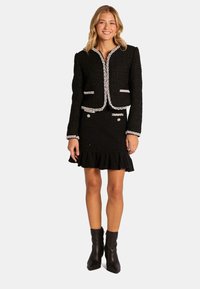 Czarna marynarka z tweedu z kontrastowymi białymi wykończeniami, w komplecie z dopasowaną, strukturalną spódnicą z falbaną u dołu i ozdobnymi guzikami.