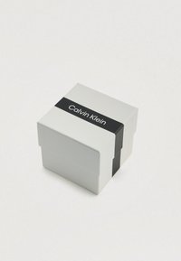 Boîte en carton blanche avec une bande noire portant le logo "Calvin Klein". Forme carrée avec un couvercle et sans motifs ni textures visibles.