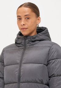 Giacca imbottita grigia con cappuccio, caratterizzata da una zip frontale, sezioni trapuntate e una texture liscia. Include dettagli hardware regolabili.