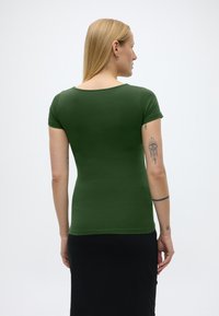 Blondýna s tetováním na pažích, oblečená v přiléhavém zeleném tričku s krátkým rukávem a černé sukni, otočená zády k jednobarevnému světlému pozadí.