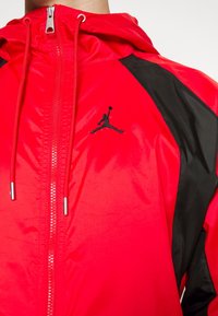 Veste légère rouge avec un panneau latéral noir, fermeture éclair et capuche ajustable. Présente un logo brodé noir sur la poitrine.