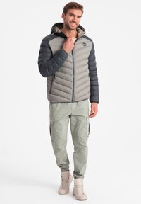 Vaatteena on harmaa ja vaaleanharmaa pehmustettu takki hupulla, jossa on vetoketjukiinnitys ja vinorahasysteemi. Yhdistetty vihreisiin cargo-housuihin ja beigeihin saappaisiin.