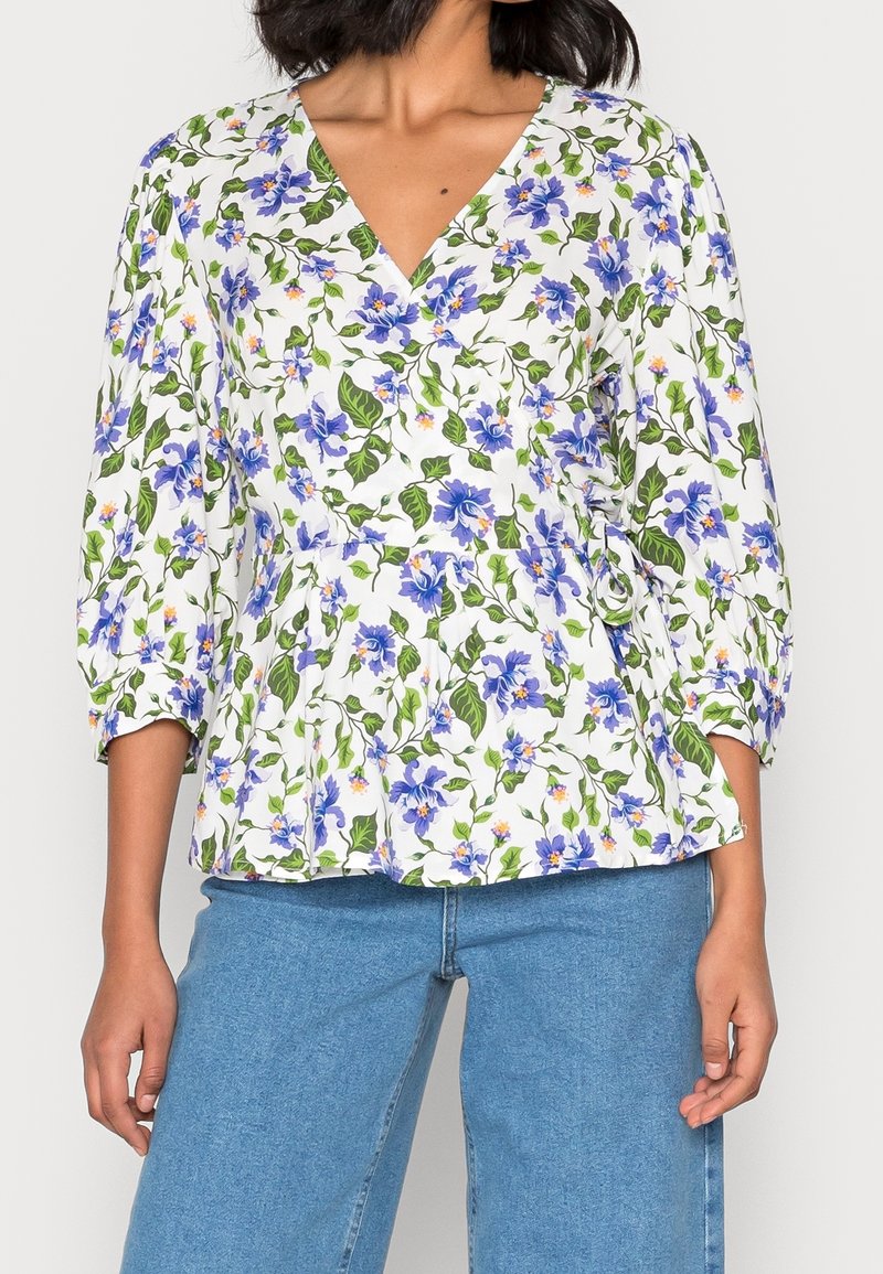 Top avvolgente bianco con fiori blu e foglie verdi, maniche a sbuffo lunghe, scollatura a V e un dettaglio arricciato in vita.