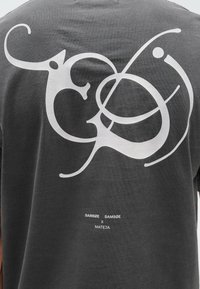 Camiseta gris oscuro con un gran diseño abstracto blanco en la parte posterior y una pequeña marca de texto situada debajo. Material de algodón.