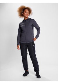 Tuta sportiva di colore grigio scuro con felpa con zip e pantaloni neri con una striscia a pied-de-poule. Realizzata in materiale liscio con loghi.