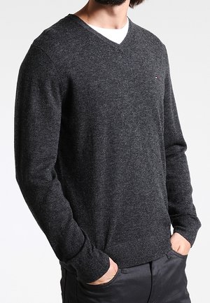 Mężczyzna ubrany w ciemnoszary sweter z dekoltem w serek na białą koszulę, z rękami w kieszeniach czarnych spodni, na białym tle.