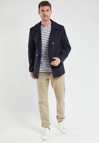 Cappotto peacoat doppiopetto blu navy con grandi bottoni. Indossato sopra una camicia bianca a righe blu navy, abbinato a pantaloni beige e sneakers bianche.