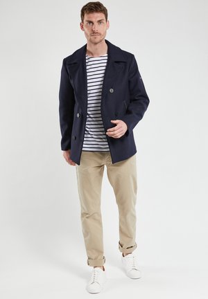 Navy double-breasted peacoat with large buttons. Worn over a white and navy striped shirt, paired with beige pants and white sneakers.