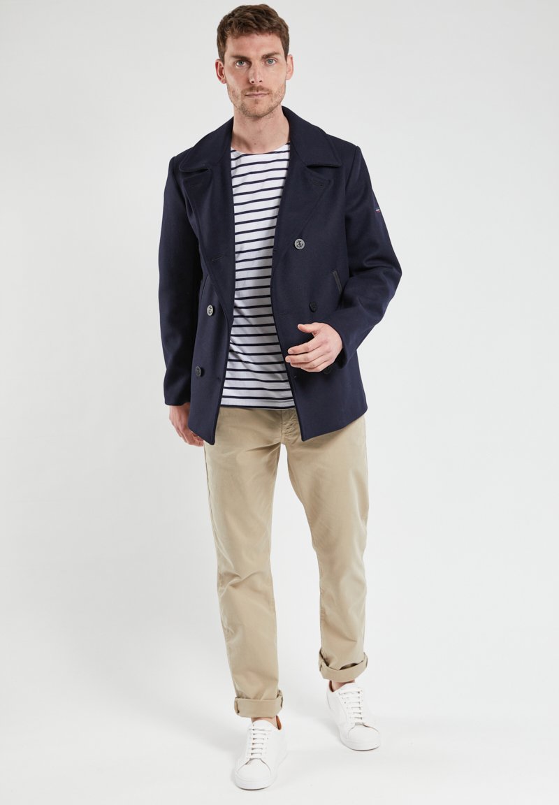 Cappotto peacoat doppiopetto blu navy con grandi bottoni. Indossato sopra una camicia bianca a righe blu navy, abbinato a pantaloni beige e sneakers bianche.