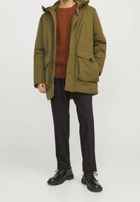 Parka de color verde oliva con capucha, que cuenta con bolsillos abotonados y cremalleras, colocada sobre un suéter texturizado color óxido y pantalones oscuros, llevada con zapatos negros.