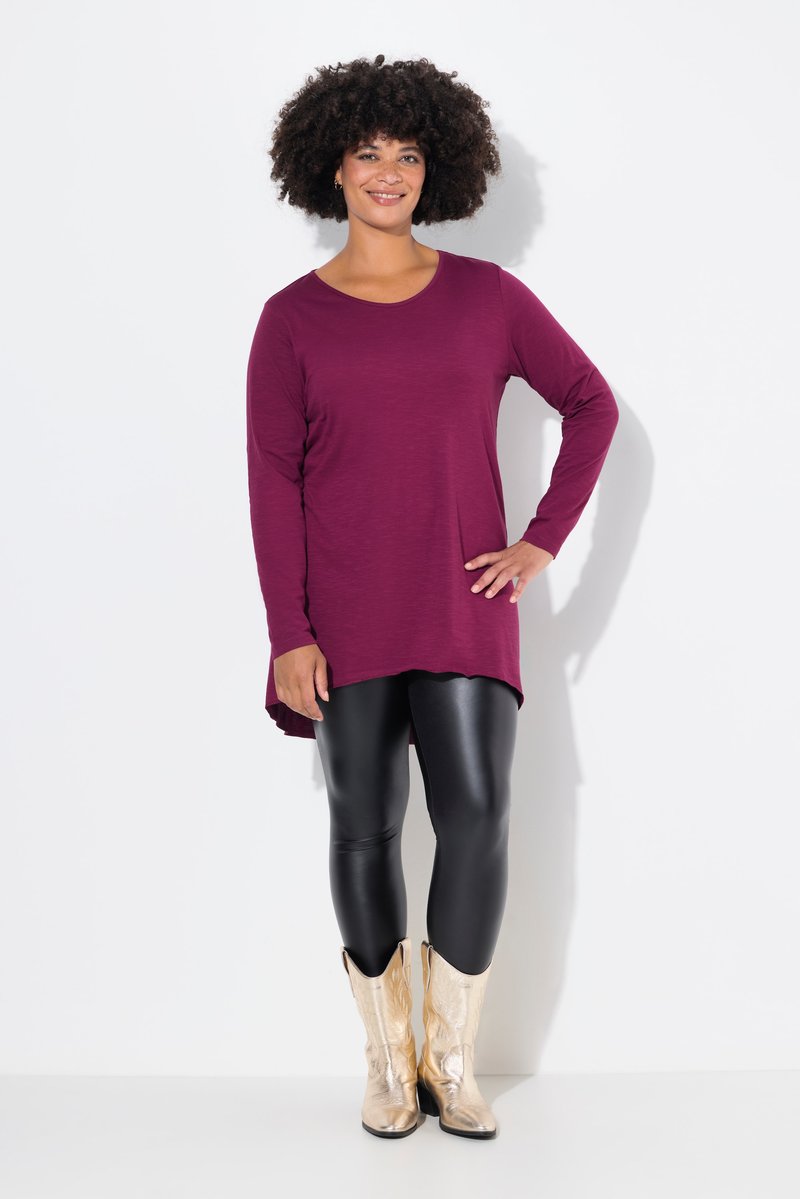 Donna in piedi con la mano sul fianco, indossa una tunica viola a maniche lunghe, leggings in pelle nera e stivali da cowboy metallici dorati su sfondo bianco.