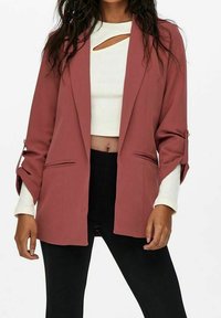 Veste rose cintrée avec deux poches avant et manches retroussées, portée sur un top blanc court et un pantalon noir.