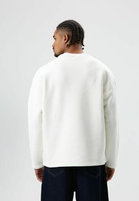 Witte oversized sweatshirt met ronde hals, doorgezakte schouders en geribbelde boorden, gecombineerd met donkere spijkerbroek. Gladde textuur en effen kleur.