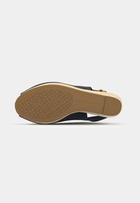 Sandal de tacón negro con suela de goma texturizada. Presenta una suela exterior de color claro y un diseño en su superficie, además de un accesorio de hardware dorado.
