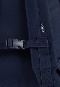 Materiál batohu námořnicky modré barvy s texturovaným povrchem; obsahuje plastovou přezku a štítek s logem označeným „POLO“. Viditelné zvýrazněné prošití.