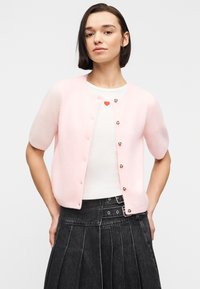 Γυναίκα φοράει ανοιχτό ροζ κοντομάνικο ζακέτα πάνω από λευκό τοπ με σχέδιο καρδιάς, συνδυασμένο με ψηλόμεση μαύρη plisés φούστα.