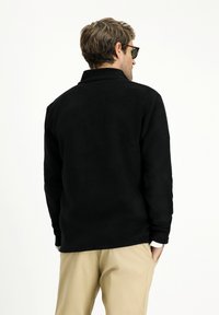 Sudadera de forro polar negra con cuello, con mangas largas, tejido texturizado y un corte relajado. Llevada con pantalones beiges.