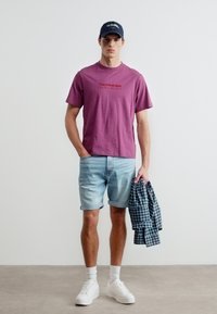 Νέος άνδρας που φοράει μωβ μπλουζάκι με τύπωμα, ανοιχτόχρωμα τζιν σορτς, λευκά αθλητικά παπούτσια και σκοτεινό μπλε καπέλο, κρατώντας ένα διπλωμένο πουκάμισο με καρό σχέδιο.