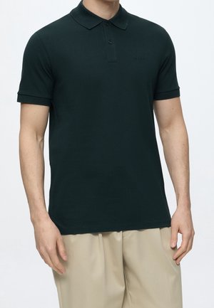 Man wearing a dark green short-sleeve polo shirt with beige pants, standing against a plain light background.