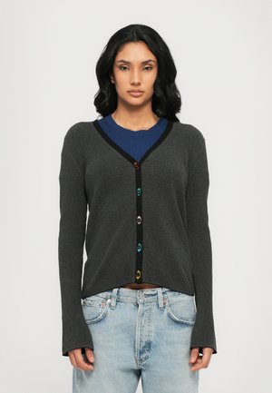 Grijze geribde cardigan met lange mouwen, zwarte accenten en kleurrijke knopen. G gedragen over een blauwe top met textuurdetails en losse, lichte spijkerbroek.