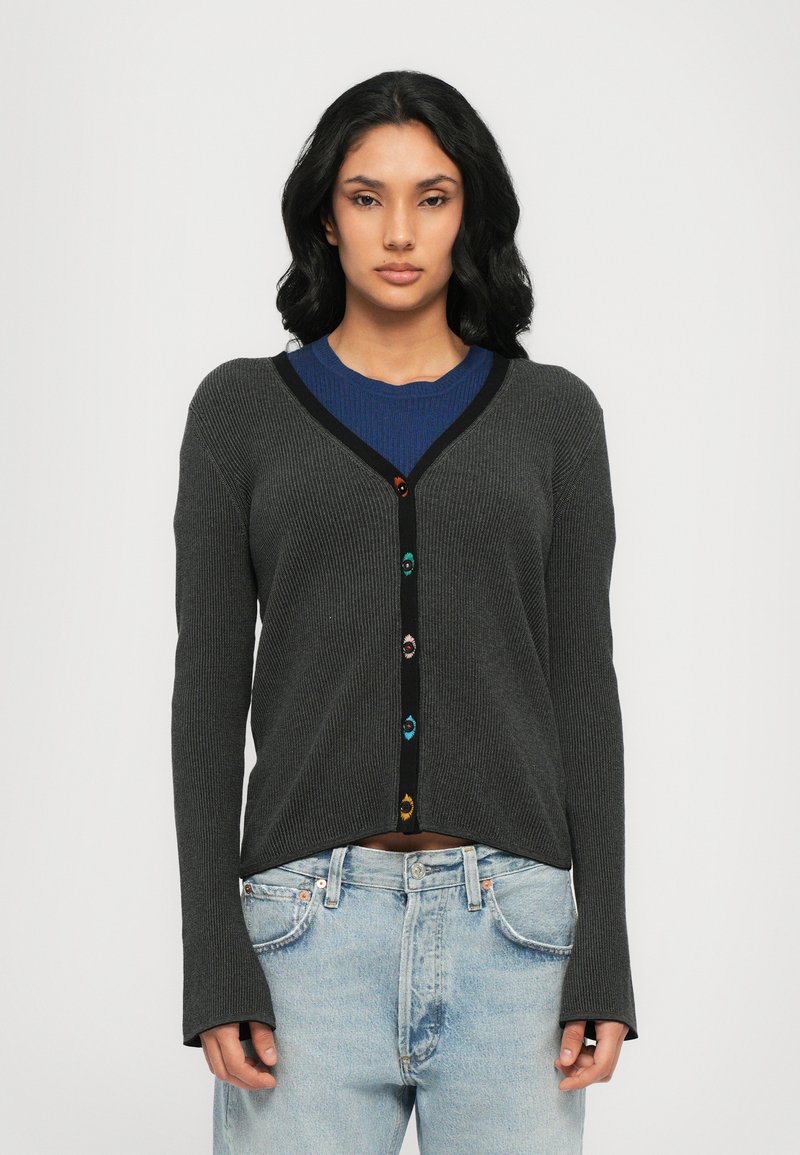 Cardigan grigio a coste con maniche lunghe, rifiniture nere e bottoni colorati. Indossato sopra un top blu con dettagli testurizzati e jeans chiari dalla vestibilità ampia.