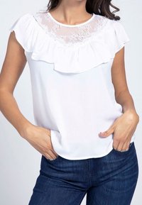 Bílá blůza s volánkovým výstřihem a krajkovým vsadkou. Krátké, splývavé rukávy. Měkká látka se volně převaluje přes uvolněný střih, doplněná tmavými džíny.