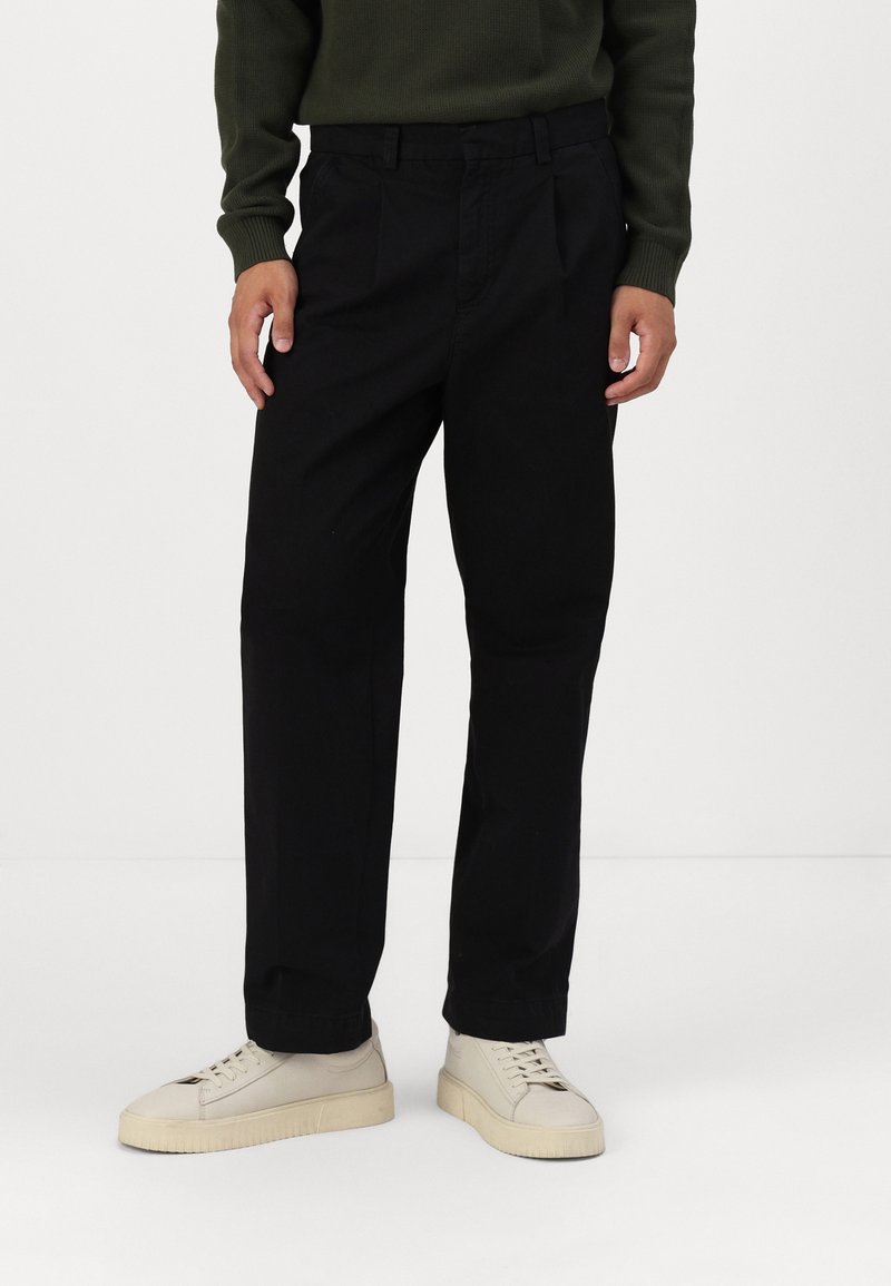 Svarta bomullsbyxor med rak bendesign, prydda med en framficka och bälteshällor, i kombination med beige sneakers.