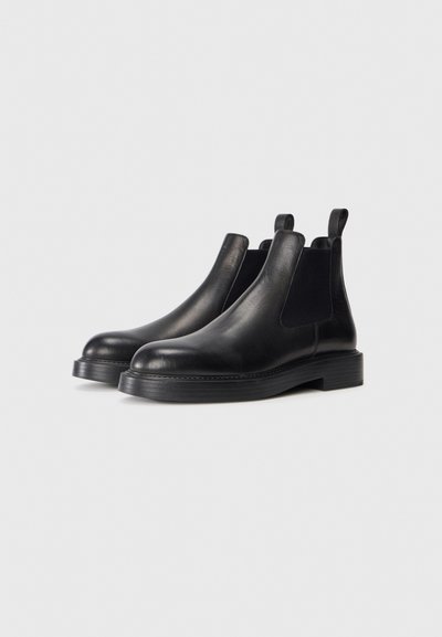 Svarta läder Chelsea-boots med elastiska sidopaneler och dragflikar, med rundade tår och tjocka låga klackar på en vit bakgrund.