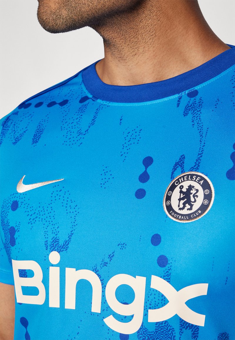 Niebieska koszulka piłkarska z okrągłym kołnierzykiem, teksturowanym wzorem, złotym logo Nike oraz herbem Chelsea FC na piersi. Na koszulce wyświetlone białe logo "Bingx".