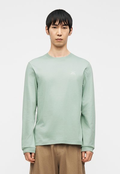 Άνδρας που φορά ανοιχτό πράσινο μακρυμάνικο πουκάμισο με μικρό άσπρο λογότυπο στο στήθος και μπεζ παντελόνι, στέκεται μπροστά σε απλό λευκό φόντο.