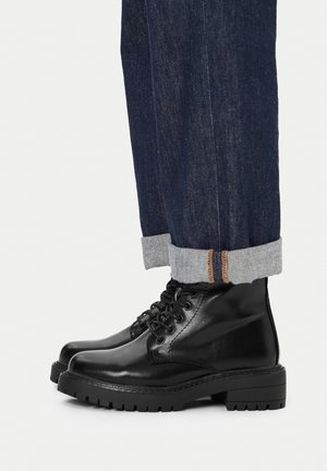 Sorte ankelstøvler i læder med runde snuder, snøredesign og robuste såler, kombineret med mørk denimjeans rullet op ved buksebenene.